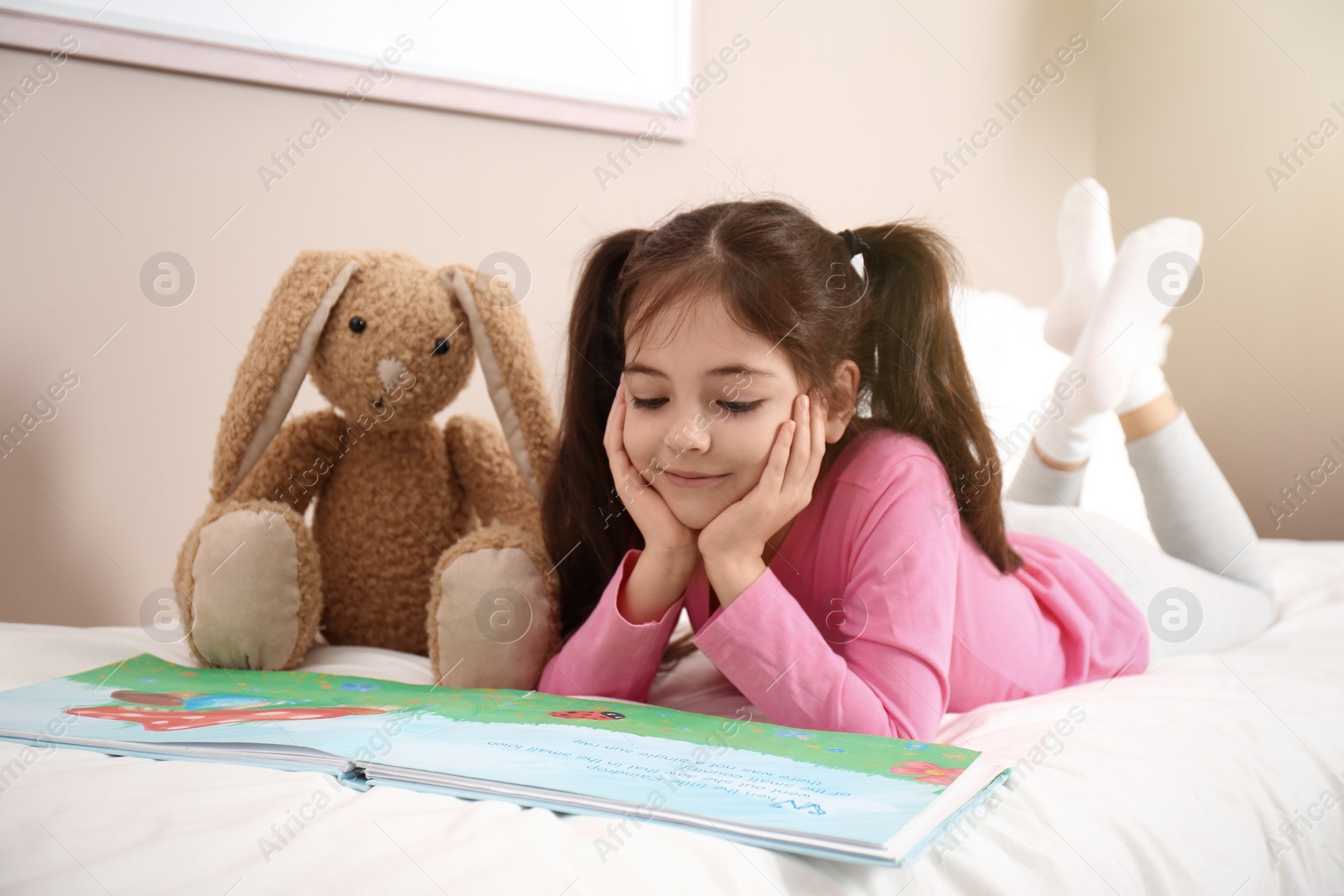 Photo of Little girl with toy bunny reading book on bed at home