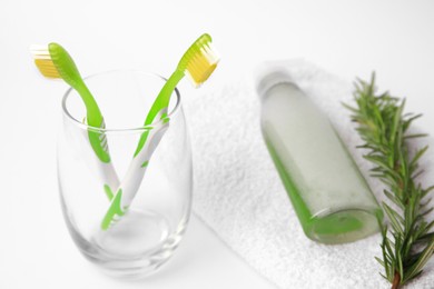 Photo of Light green toothbrushes in glass holder, terry towel, bottle of cosmetic product and rosemary on white background