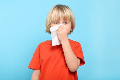 Boy blowing nose in tissue on light blue background. Cold symptoms