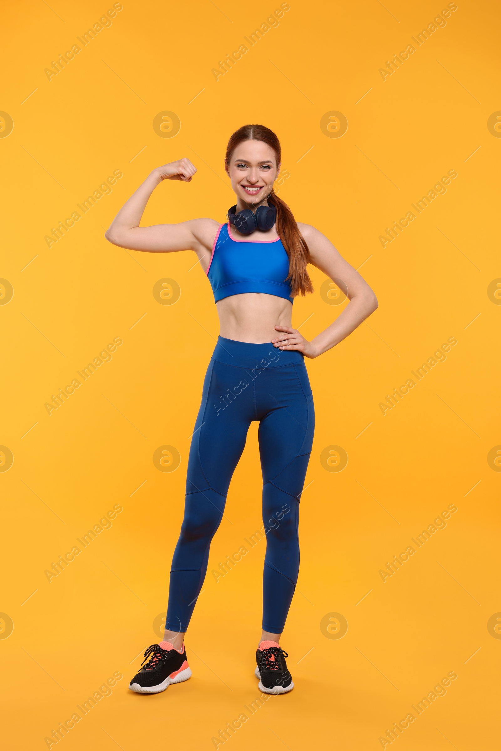 Photo of Young woman in sportswear and headphones showing muscles on yellow background