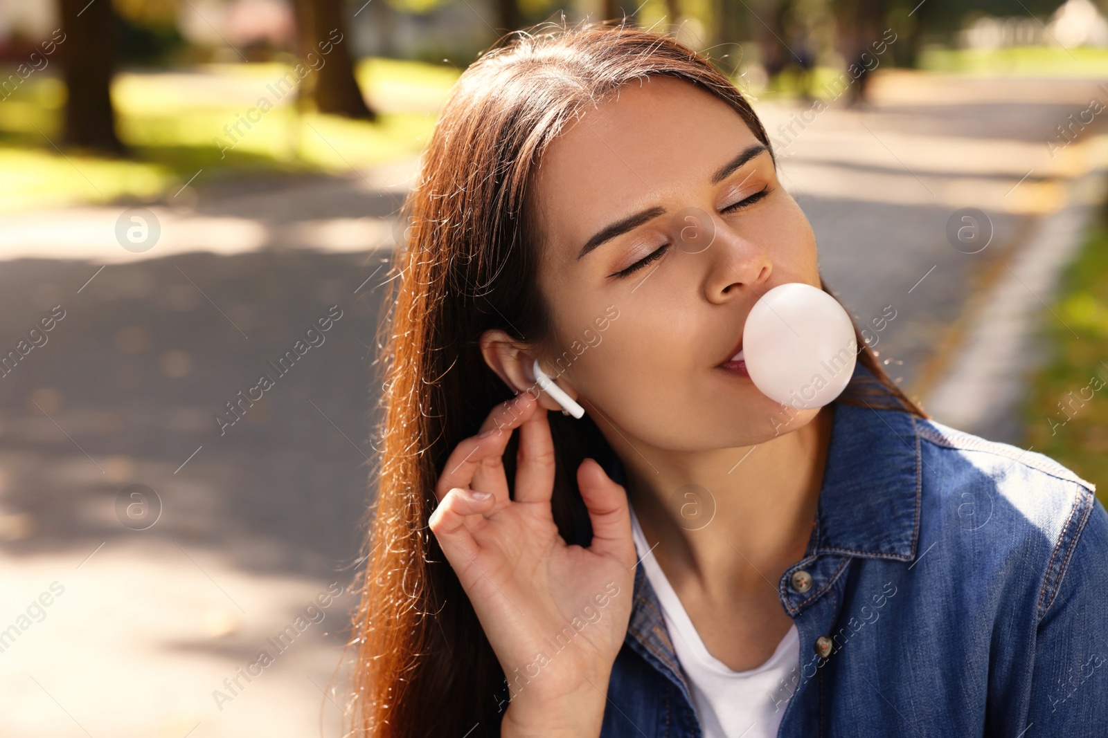 Photo of Beautiful young woman with wireless headphones blowing chewing gum outdoors, space for text