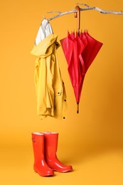 Photo of Closed red umbrella, stylish raincoat and rubber boots hanging on branch against yellow background
