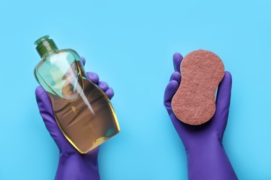 Woman in rubber gloves holding sponge and detergent on light blue background, top view