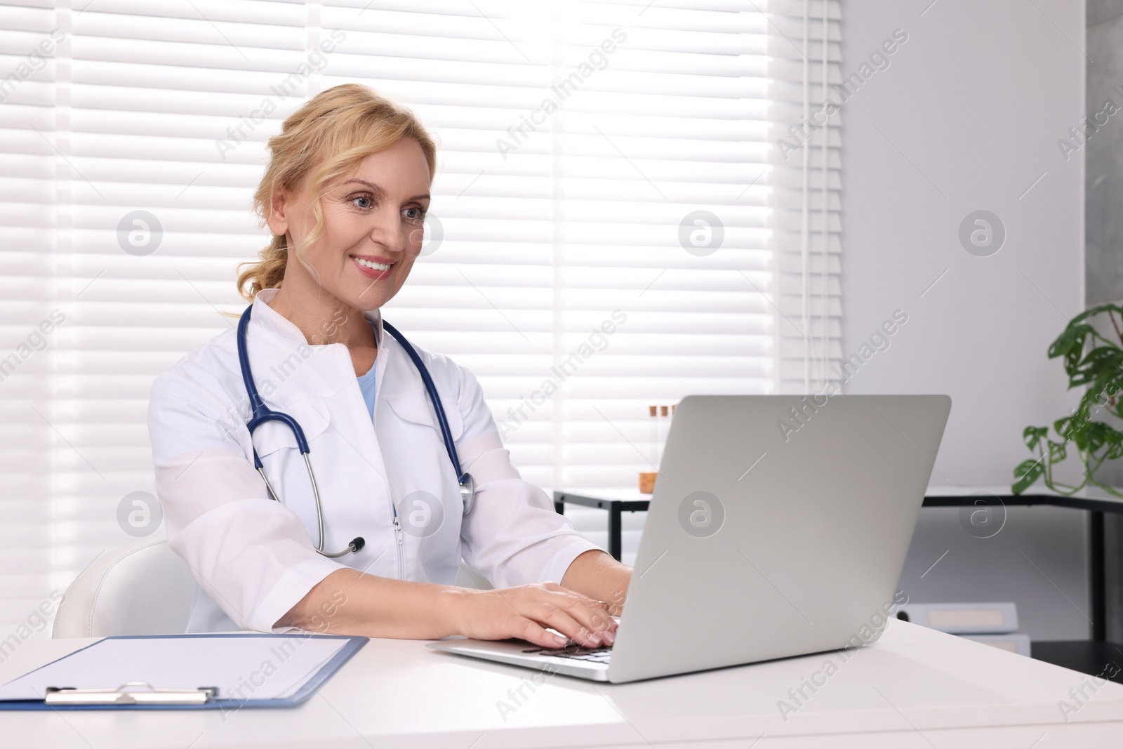 Photo of Doctor with laptop and clipboard at white table in clinic. Patient consultation