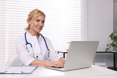 Doctor with laptop and clipboard at white table in clinic. Patient consultation