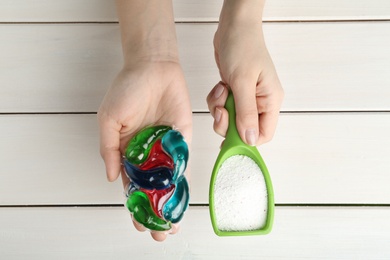 Woman holding laundry capsules and detergent powder above white wooden table, top view