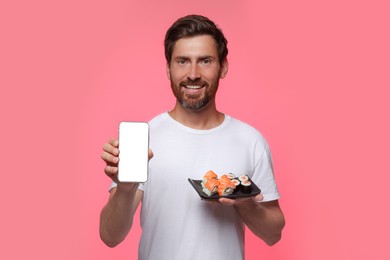 Happy man holding plate with tasty sushi rolls and smartphone against pink background