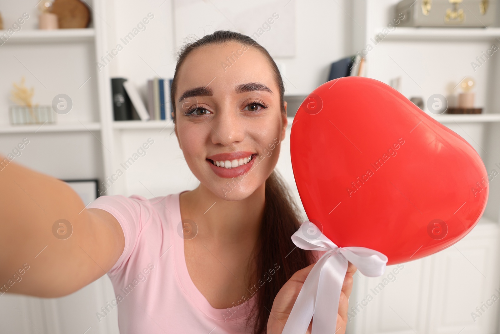 Photo of Happy young woman holding red heart shaped balloon and taking selfie at home