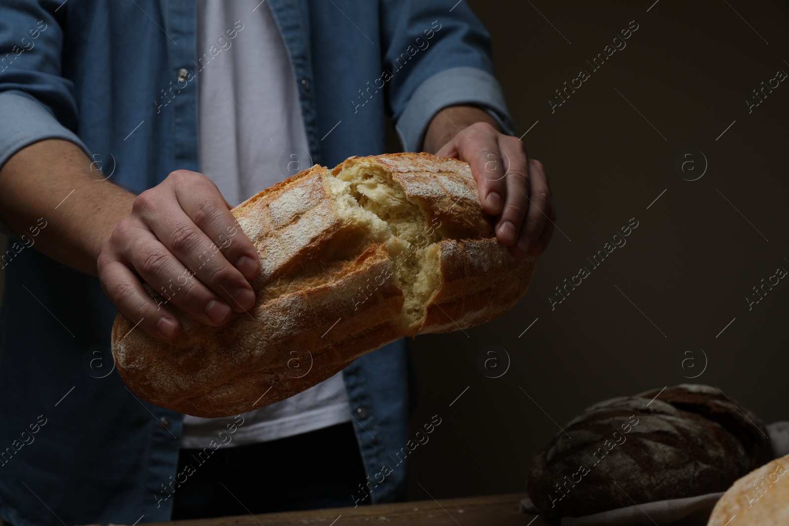 Photo of Man breaking loaf of fresh bread at wooden table, closeup