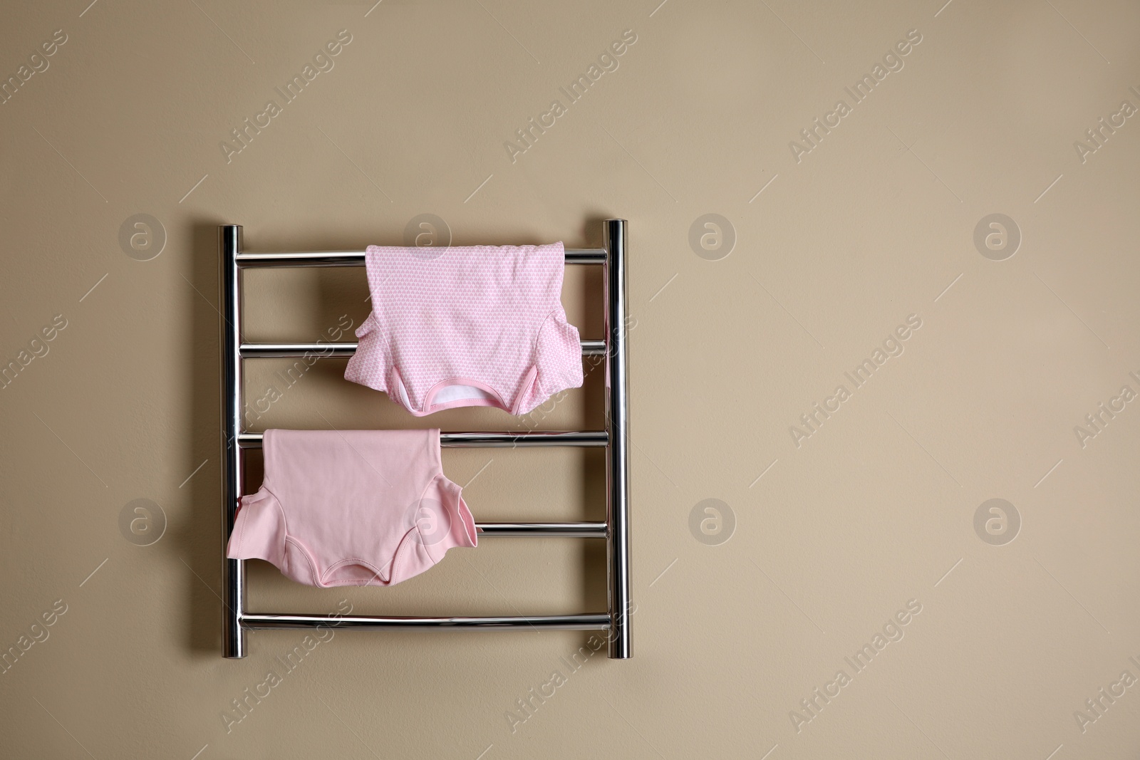 Photo of Modern heated towel rail with baby clothes on beige wall. Space for text