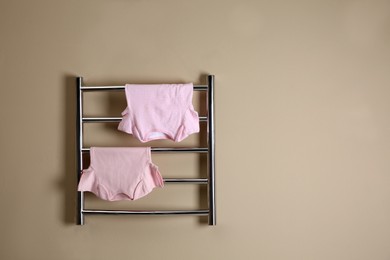 Photo of Modern heated towel rail with baby clothes on beige wall. Space for text