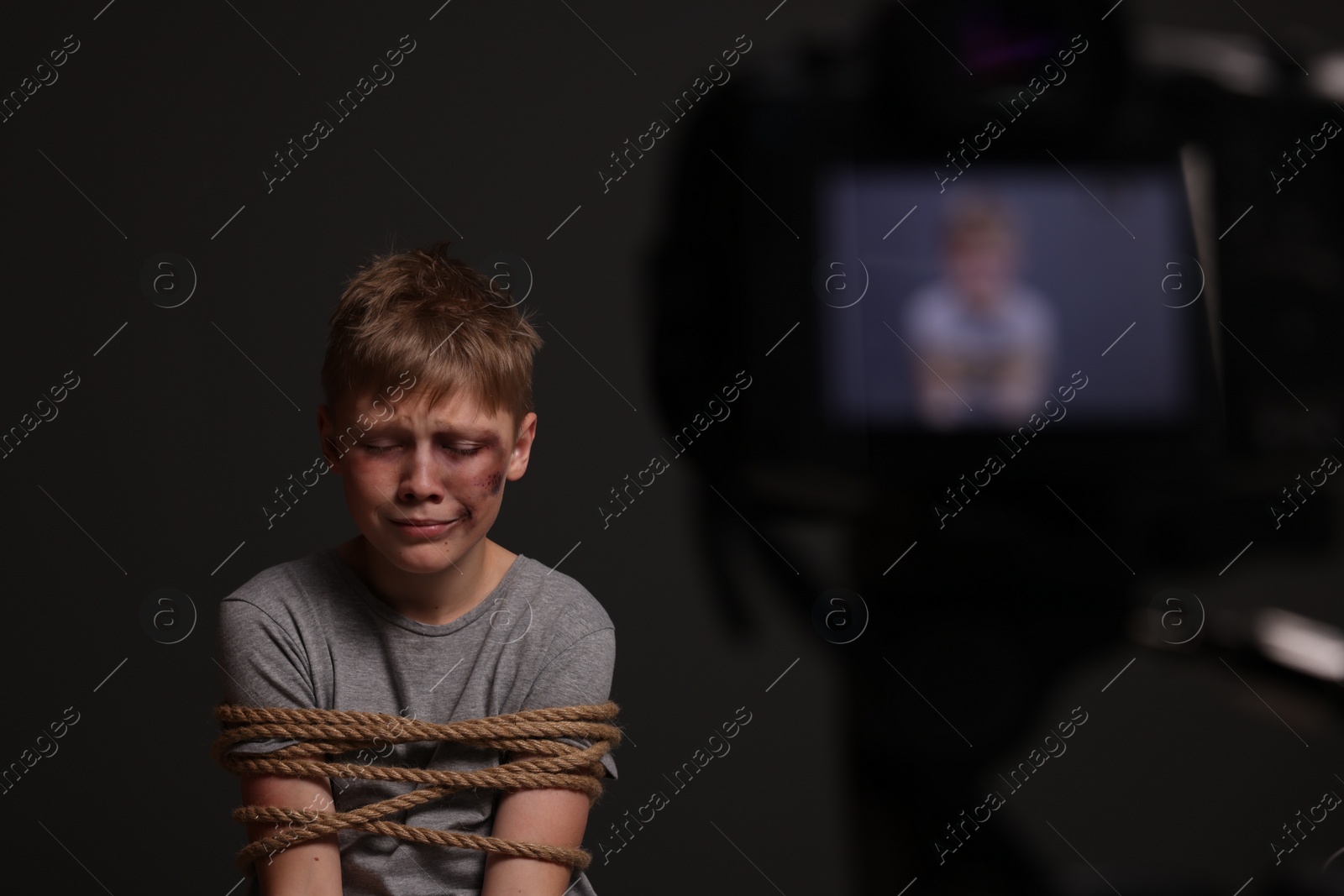 Photo of Little boy with bruises tied up and taken hostage near camera on dark background, selective focus