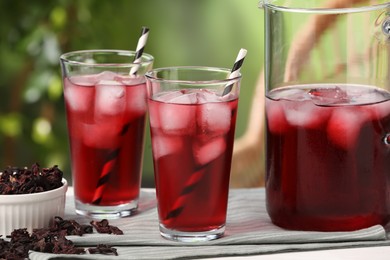Photo of Refreshing hibiscus tea with ice cubes and dry roselle flowers on white table against blurred green background