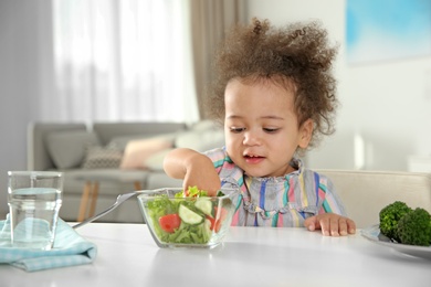 Photo of Cute African-American girl eating vegetable salad at table in room
