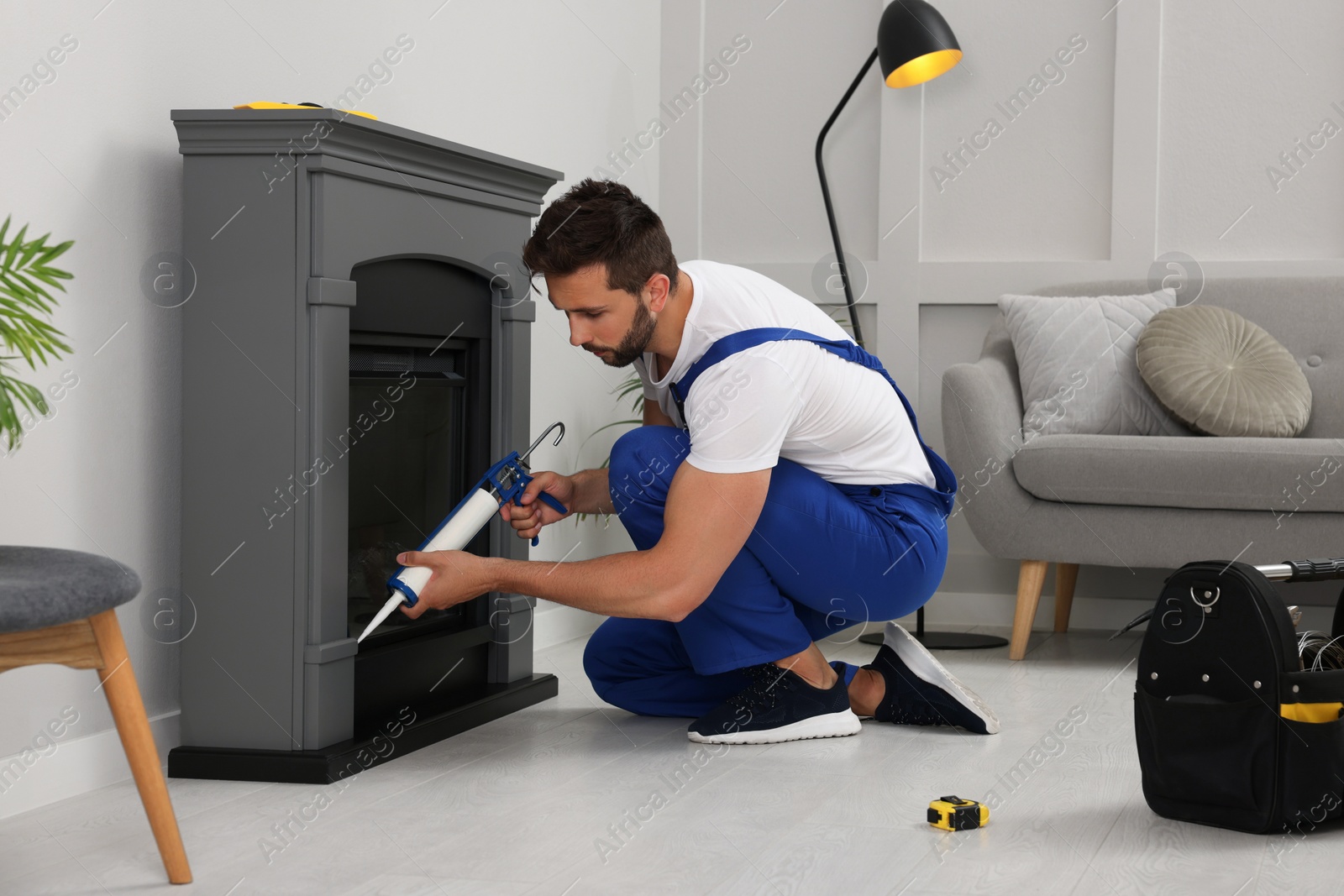 Photo of Professional technician sealing electric fireplace with caulk near white wall in room