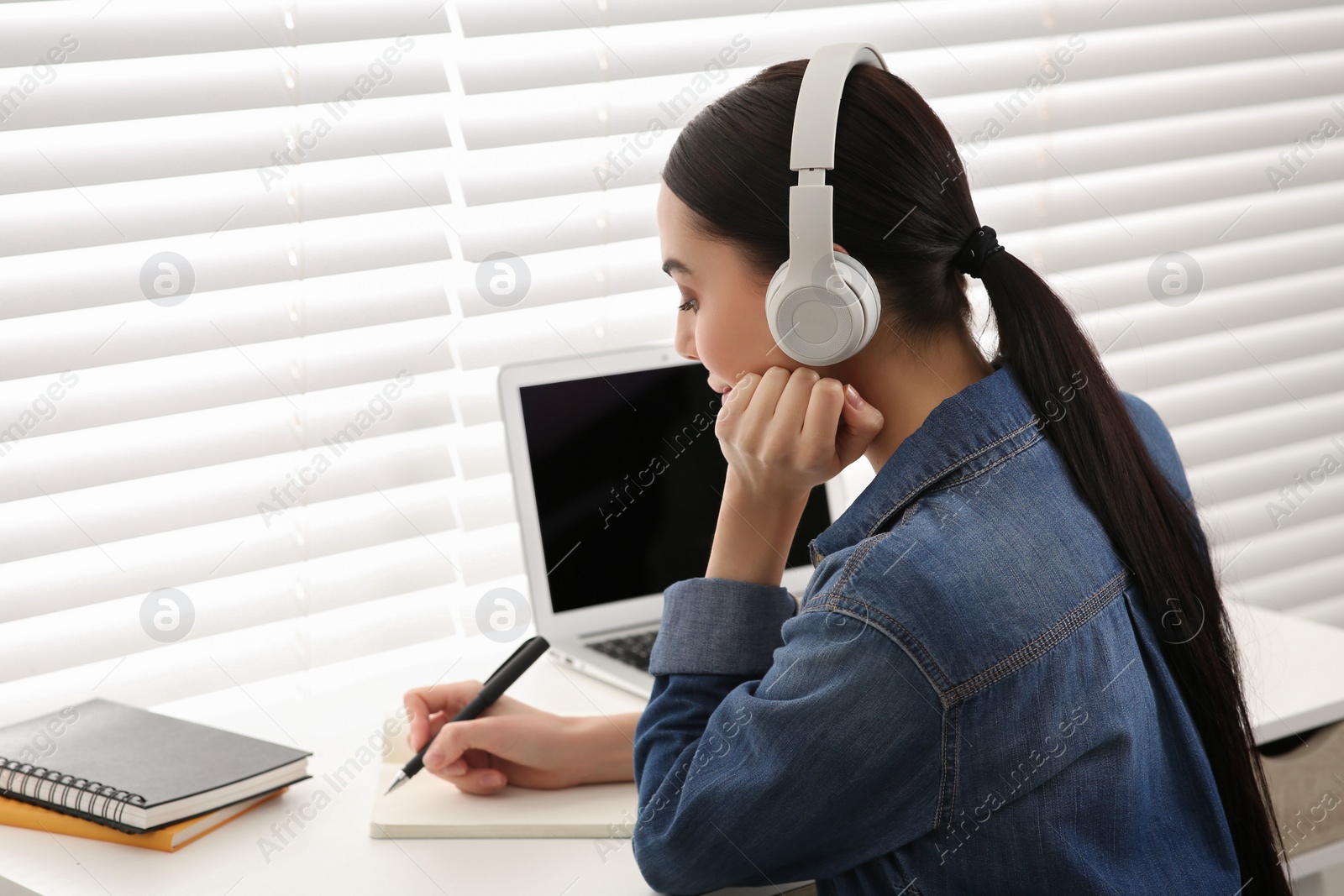 Photo of Online translation course. Student in headphones writing near laptop indoors