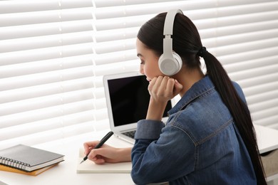 Photo of Online translation course. Student in headphones writing near laptop indoors