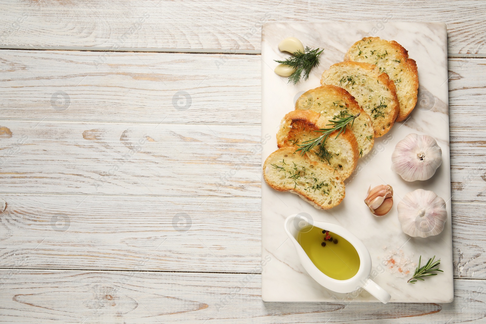 Photo of Tasty baguette with garlic, dill, rosemary and oil on white wooden table, top view. Space for text