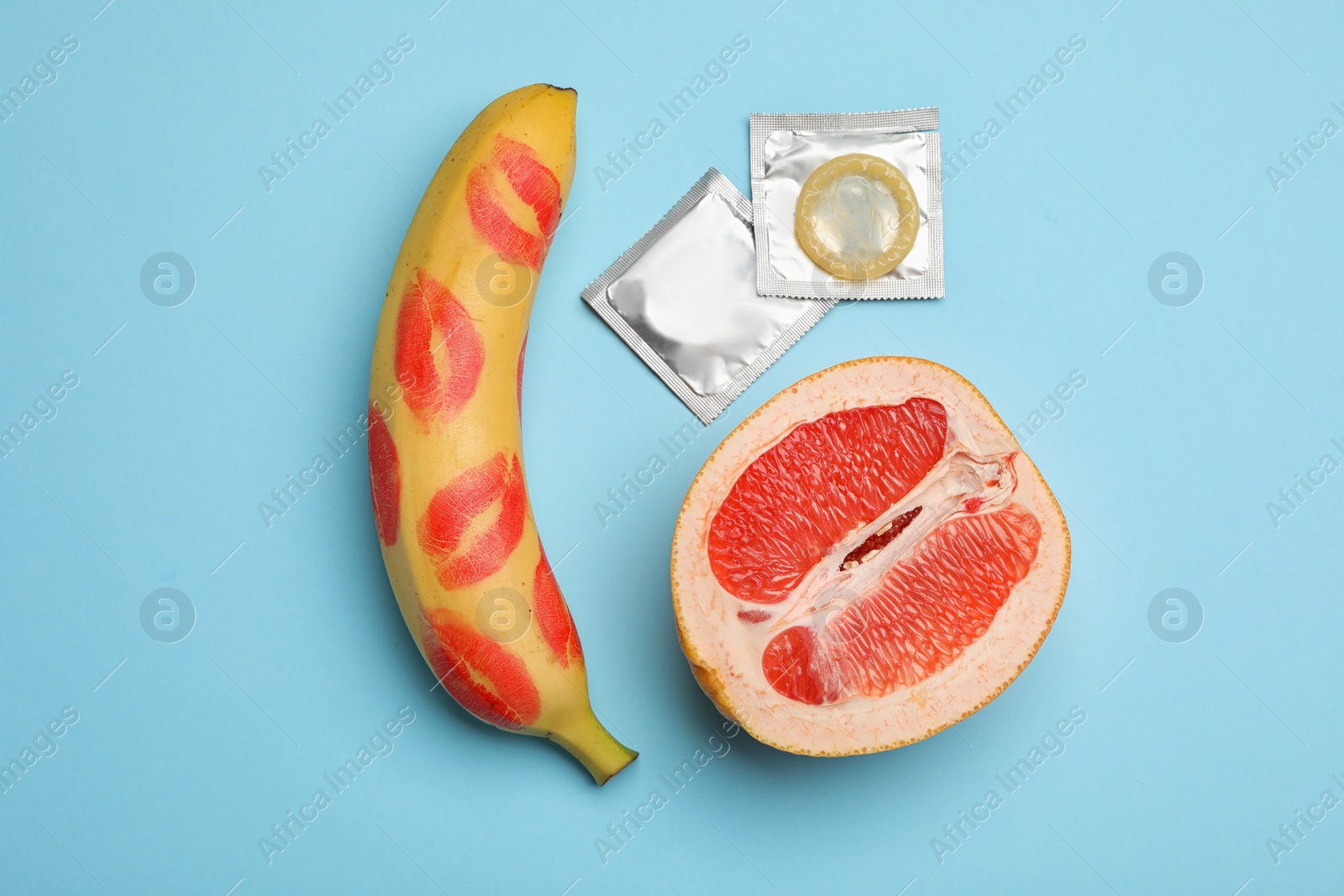 Photo of Condoms, cut grapefruit and banana with lipstick kiss marks on light blue background, flat lay. Safe sex