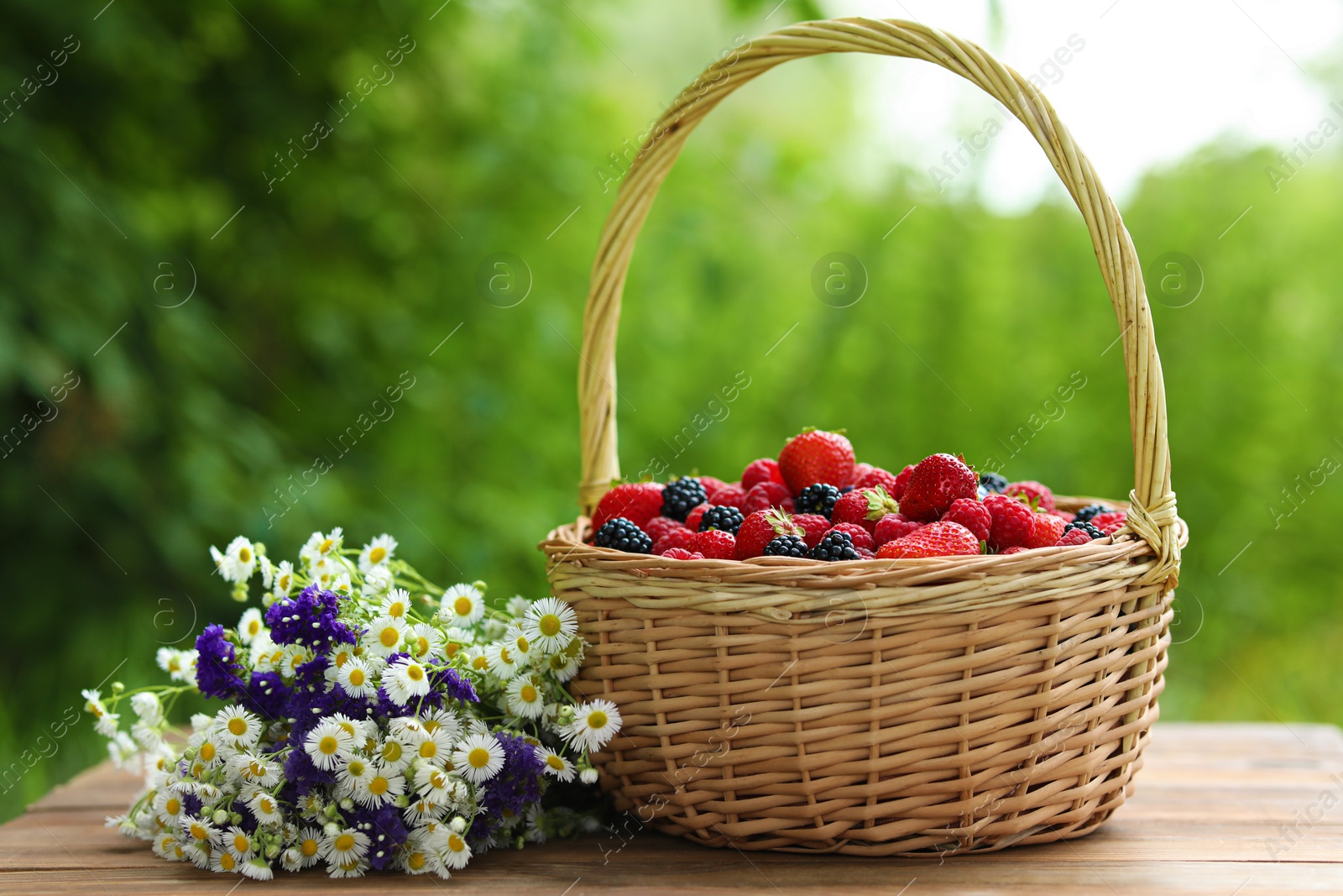 Photo of Wicker basket with different fresh ripe berries and beautiful flowers on wooden table outdoors