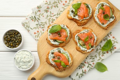 Photo of Tasty canapes with salmon, capers, cucumber and sauce on white wooden table, top view