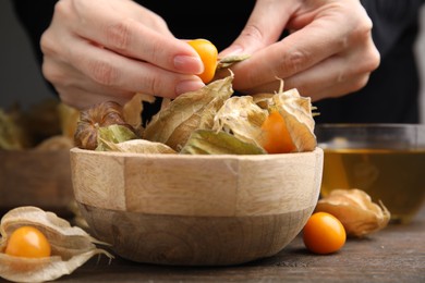 Photo of Woman peeling physalis fruit from calyxes at wooden table, closeup