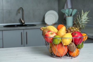 Metal basket with different ripe fruits on white wooden table in kitchen. Space for text
