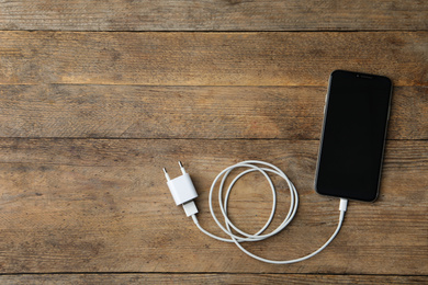 Photo of Smartphone and charging cable with adapter on wooden table, flat lay. Space for text