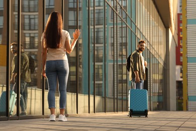Photo of Long-distance relationship. Woman waving to her boyfriend with luggage near building outdoors