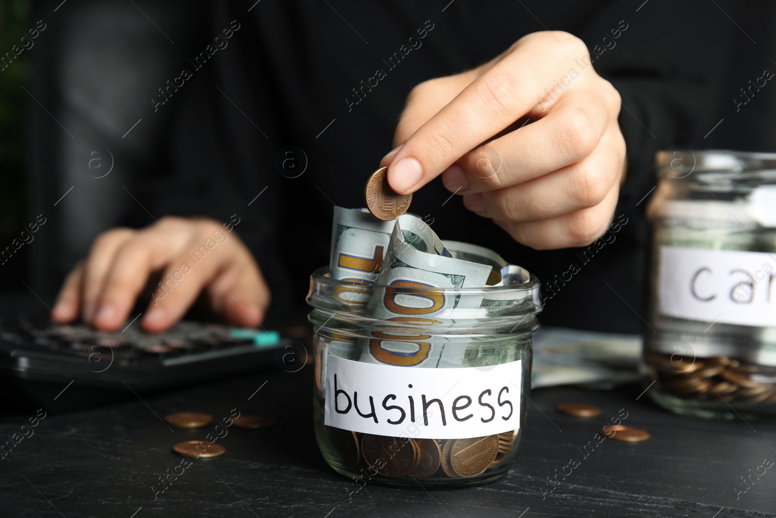 Photo of Woman putting coin into jar with money and tag BUSINESS on black table, closeup