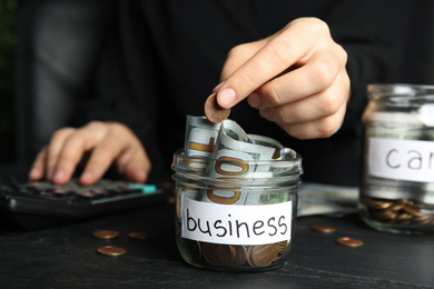 Photo of Woman putting coin into jar with money and tag BUSINESS on black table, closeup
