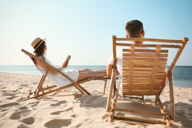 Photo of Young woman reading book and man relaxing in deck chairs on beach near sea