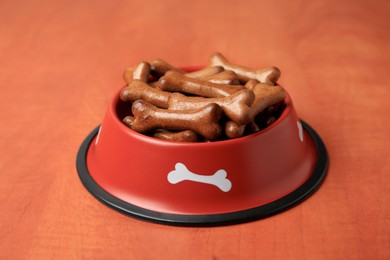 Red bowl with bone shaped dog cookies on wooden table, closeup