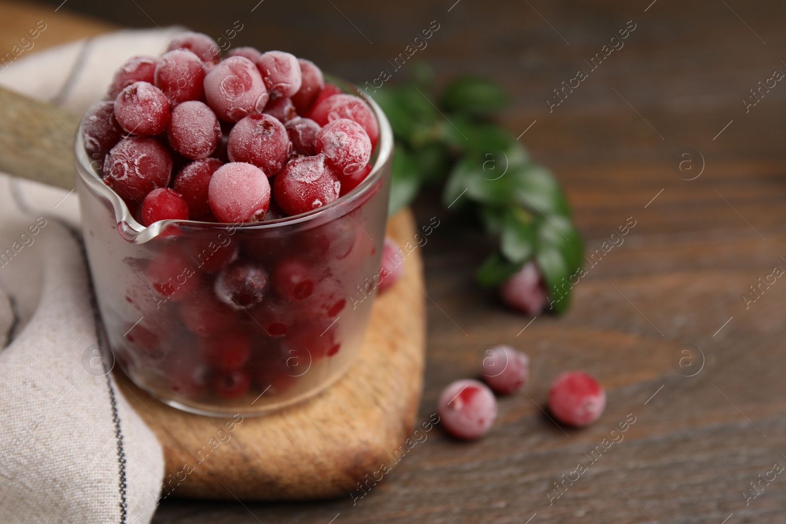 Photo of Frozen red cranberries in glass pot and green leaves on wooden table, closeup. Space for text