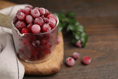 Frozen red cranberries in glass pot and green leaves on wooden table, closeup. Space for text
