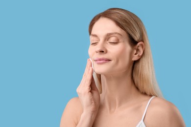 Beautiful woman removing makeup with cotton pad on light blue background, space for text