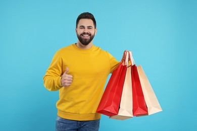 Happy man with many paper shopping bags showing thumb up on light blue background