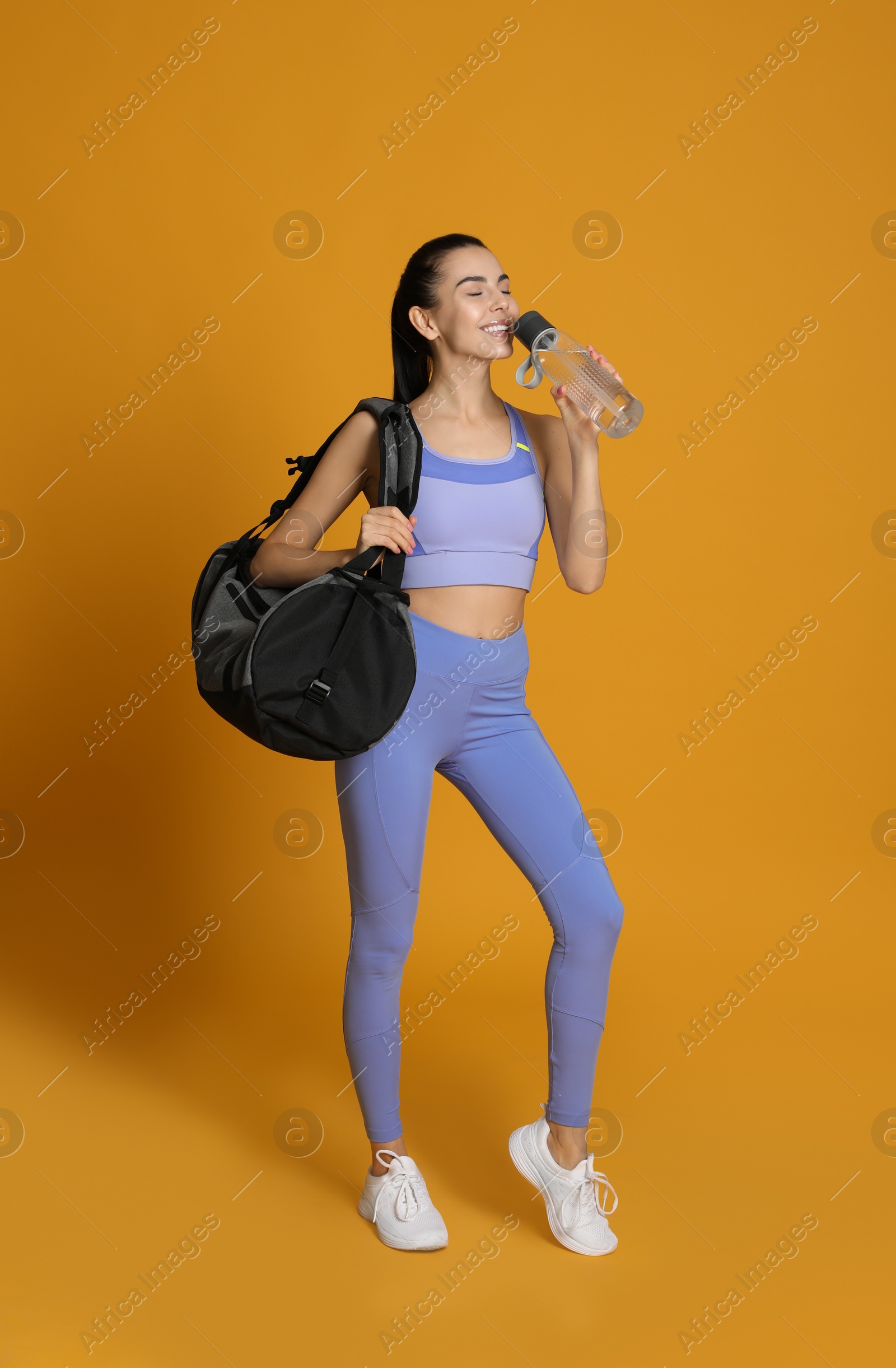 Photo of Beautiful woman with sports bag drinking water on yellow background