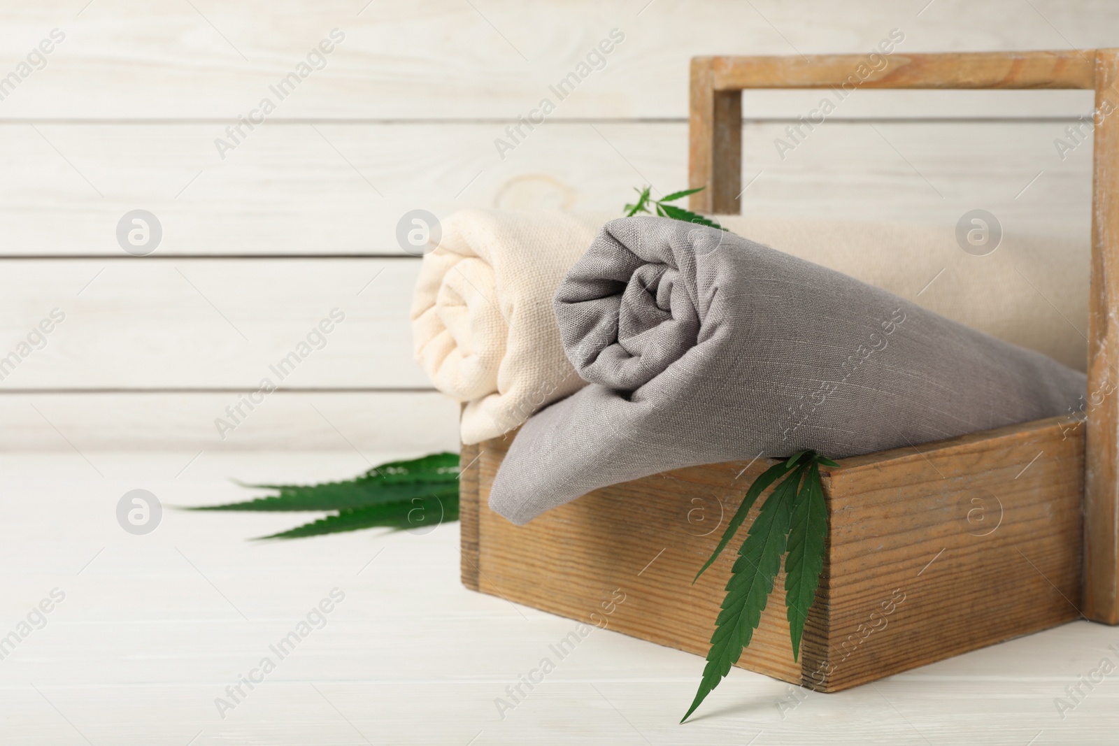 Photo of Hemp cloths in crate and green leaves on white wooden table, space for text