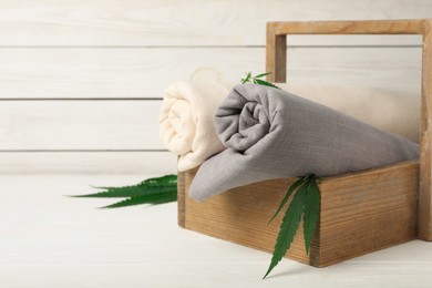 Photo of Hemp cloths in crate and green leaves on white wooden table, space for text