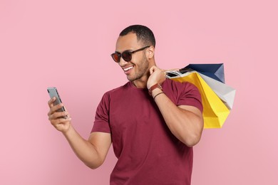 Photo of Happy African American man in sunglasses with shopping bags and smartphone on pink background