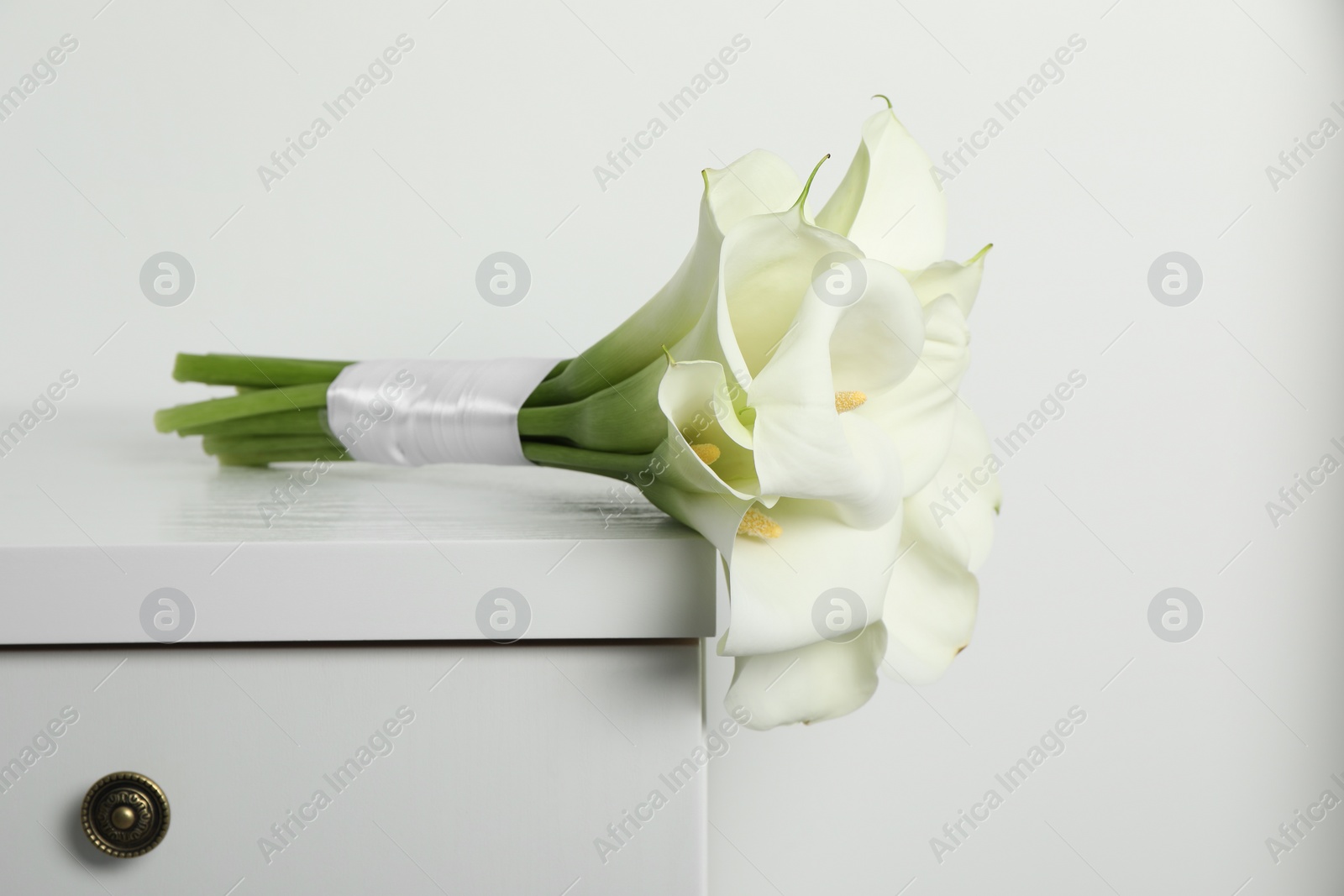 Photo of Beautiful calla lily flowers tied with ribbon on white chest of drawers indoors