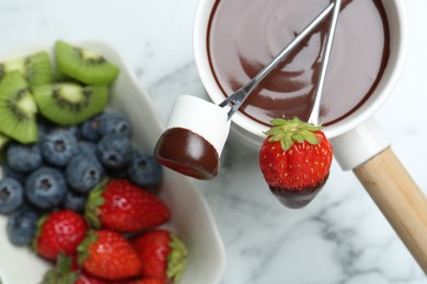 Photo of Fondue pot with melted chocolate, sweet marshmallow, fresh kiwi, different berries and forks on white marble table, flat lay