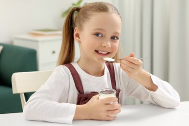 Photo of Cute little girl with tasty yogurt at white table at home