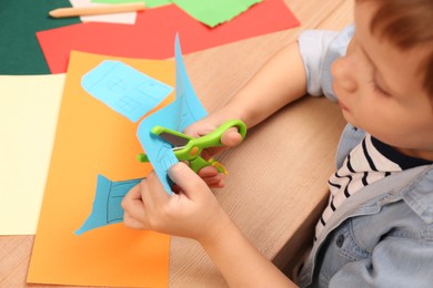 Photo of Little boy cutting color paper with scissors at table indoors, closeup