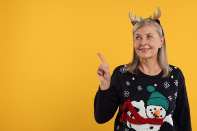 Photo of Happy senior woman in Christmas sweater and deer headband pointing at something on orange background. Space for text