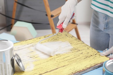 Photo of Woman using roller to paint plank with white dye at light blue wooden table indoors, closeup
