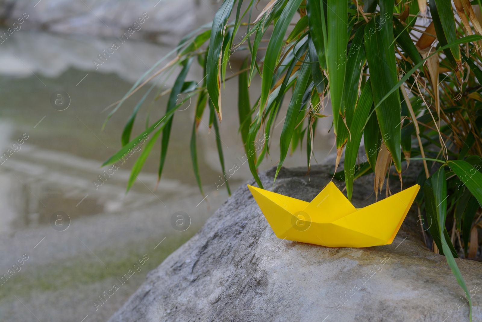 Photo of Beautiful yellow paper boat on stone and green plant leaves outdoors, space for text