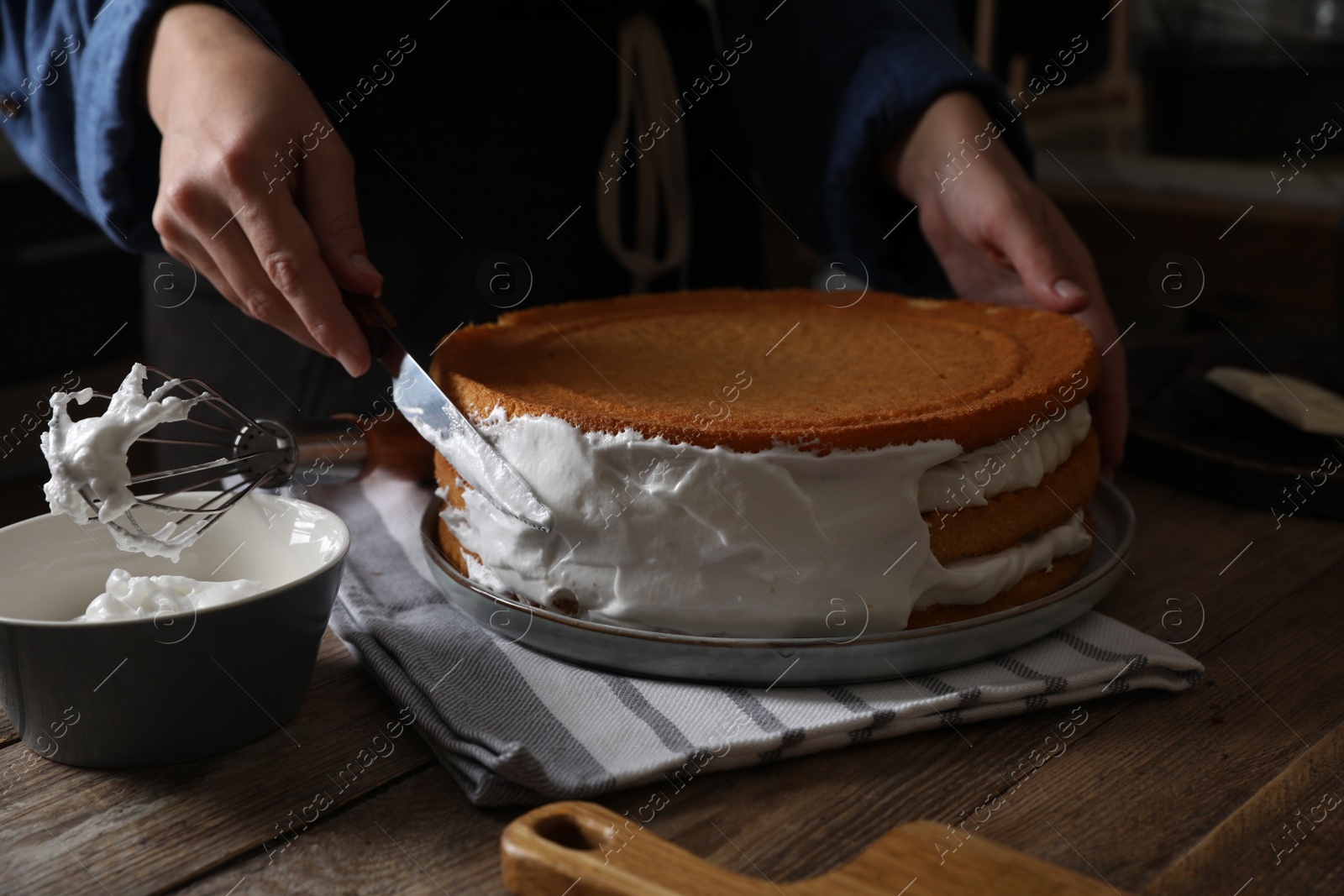 Photo of Woman smearing sides of sponge cake with cream at wooden table, closeup