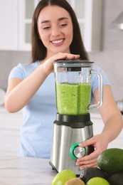 Beautiful young woman preparing tasty smoothie at white table in kitchen, focus on blender